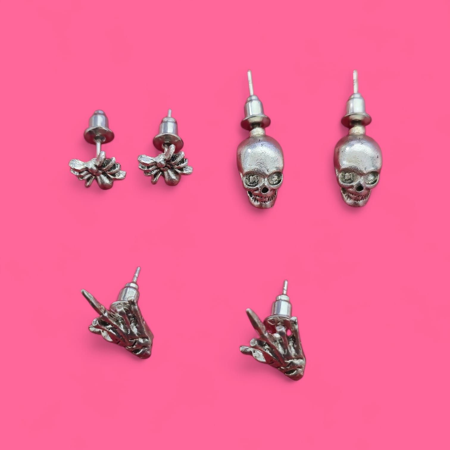 Pendientes de plata en varios diseños: floral, calavera y gesto de mano, sobre fondo rosa.