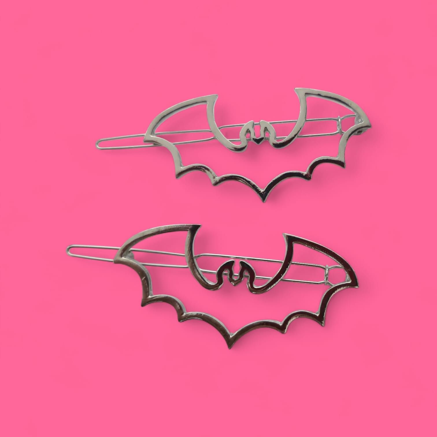 Dos pinzas para el pelo plateadas con forma de murciélago sobre un fondo rosa brillante.