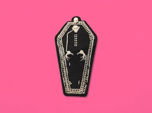 Parche negro con forma de ataúd con un vampiro en su interior sobre un fondo rosa.