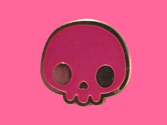 Pin de esmalte rosa con forma de calavera y ribete dorado sobre fondo rosa.