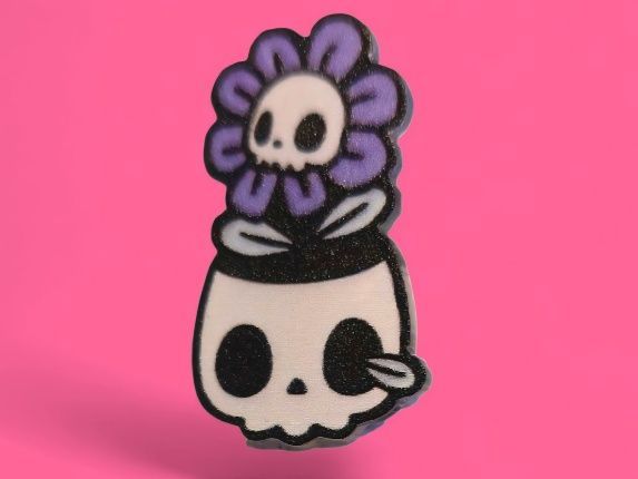 Maceta con forma de calavera con flor de calavera, pétalos de color morado, detalles en blanco y negro, sobre fondo rosa.