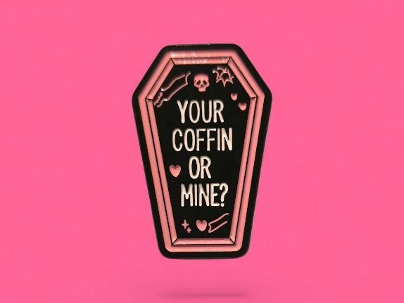 Pin en forma de ataúd de color oro rosa con "¿TU ATAÚD O EL MÍO?" sobre fondo rosa.