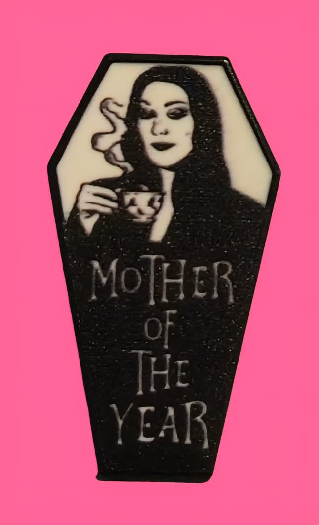 Morticia Addams en un marco con forma de ataúd, tomando té. Retrato en blanco y negro con el texto "Madre del Año".