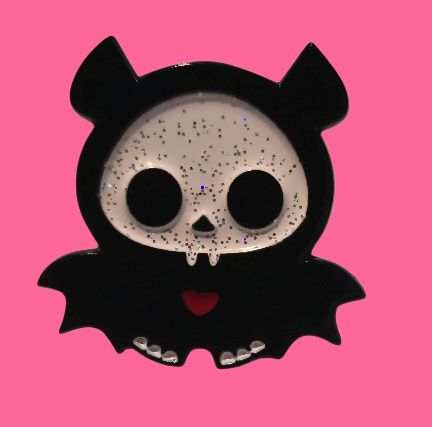 Lindo pin con forma de murciélago con cara de calavera, capa con capucha negra y corazón rojo sobre un fondo rosa.