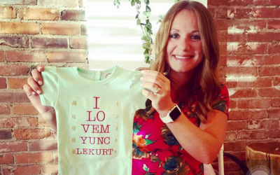 Woman holding a mint green T-shirt with red text in front of a brick wall and window blinds