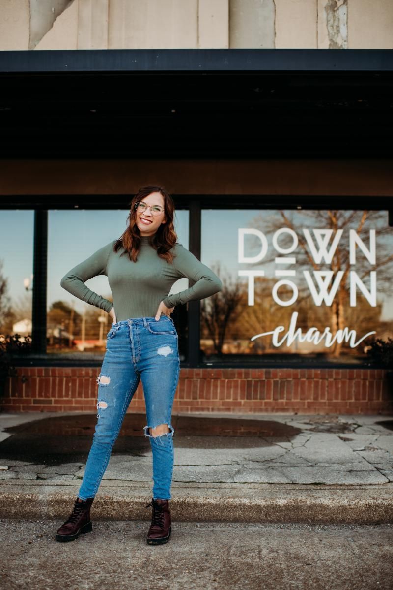 Woman standing outside a storefront with “DOWNTOWN charm” signage, hands on hips, wearing a green top and jeans.