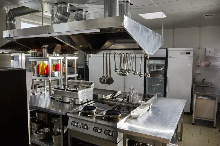 Commercial kitchen with stainless steel stoves, ventilation hood, and hanging cookware.