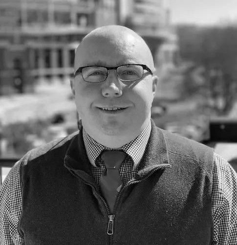 A bald man wearing glasses and a vest is smiling in a black and white photo.