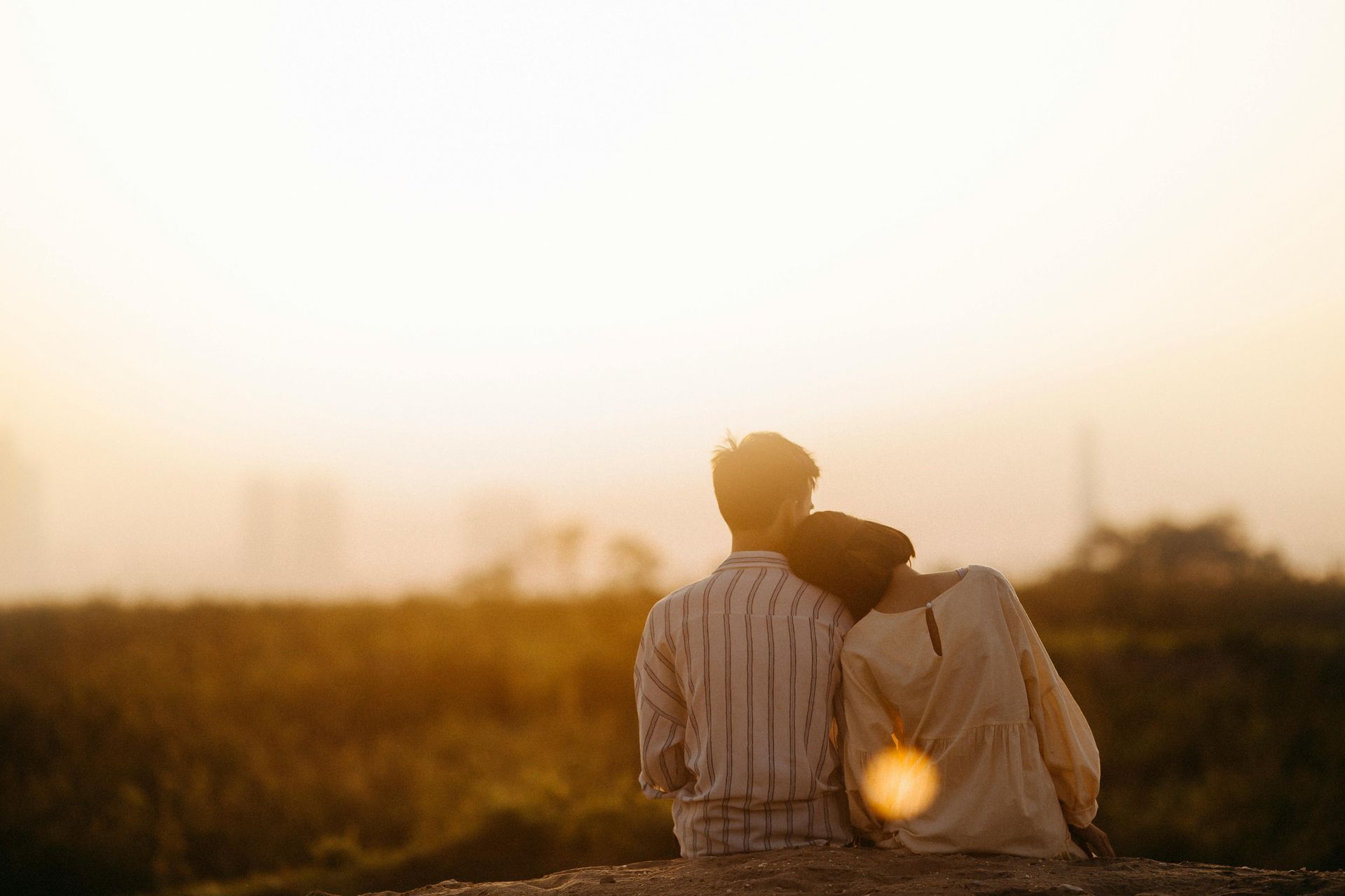 A man and a woman are sitting on top of a hill watching the sunset.
