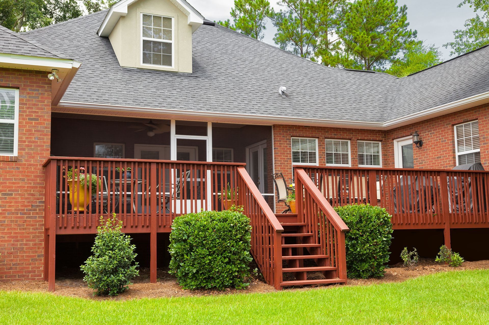 Brick house with a covered wooden deck, stairs, and green lawn under trees Brick house with a covered wooden deck, stairs, and green lawn under trees