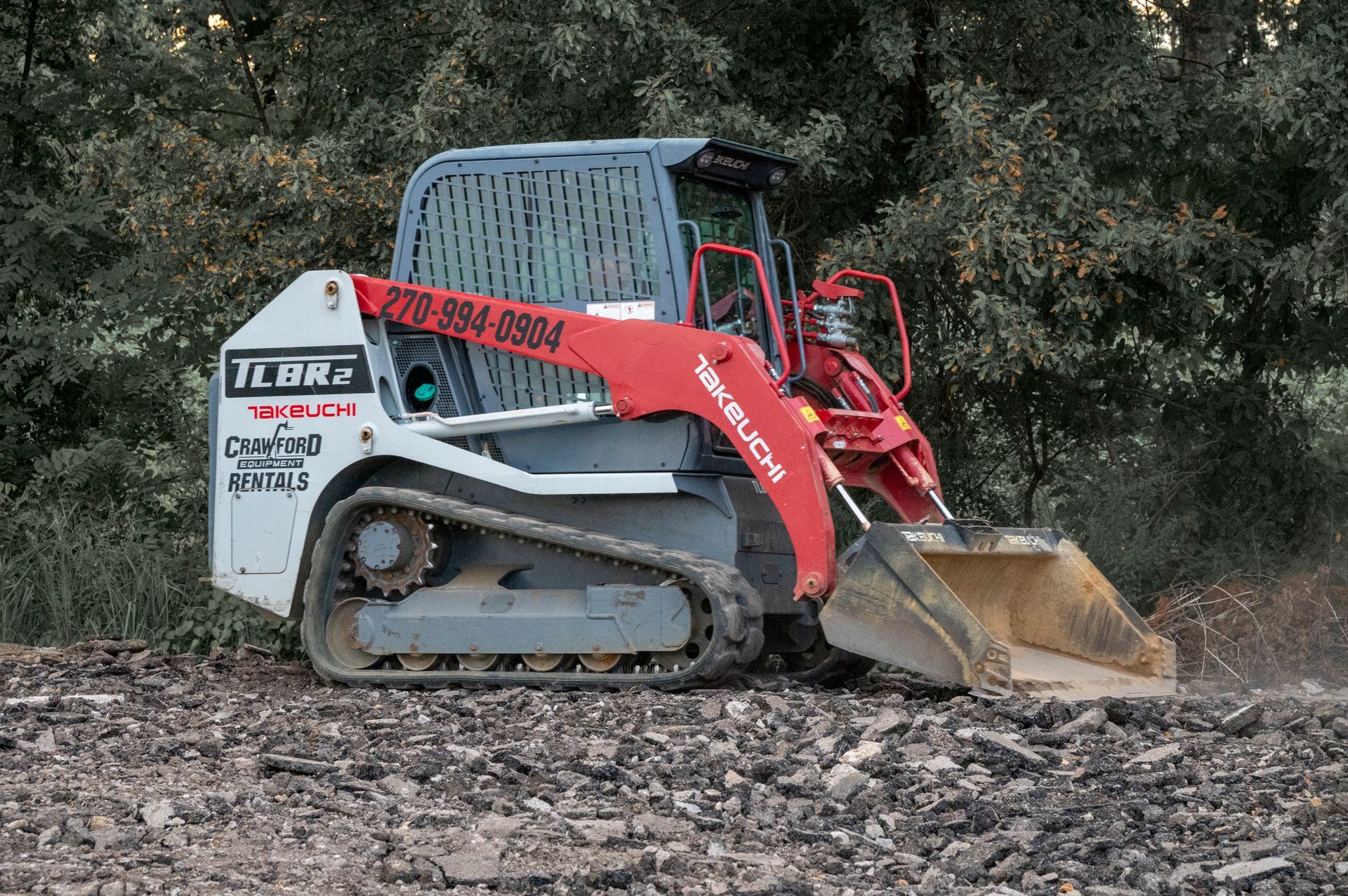 White and red Takeuchi track skid steer on rocky ground, with a forest background.