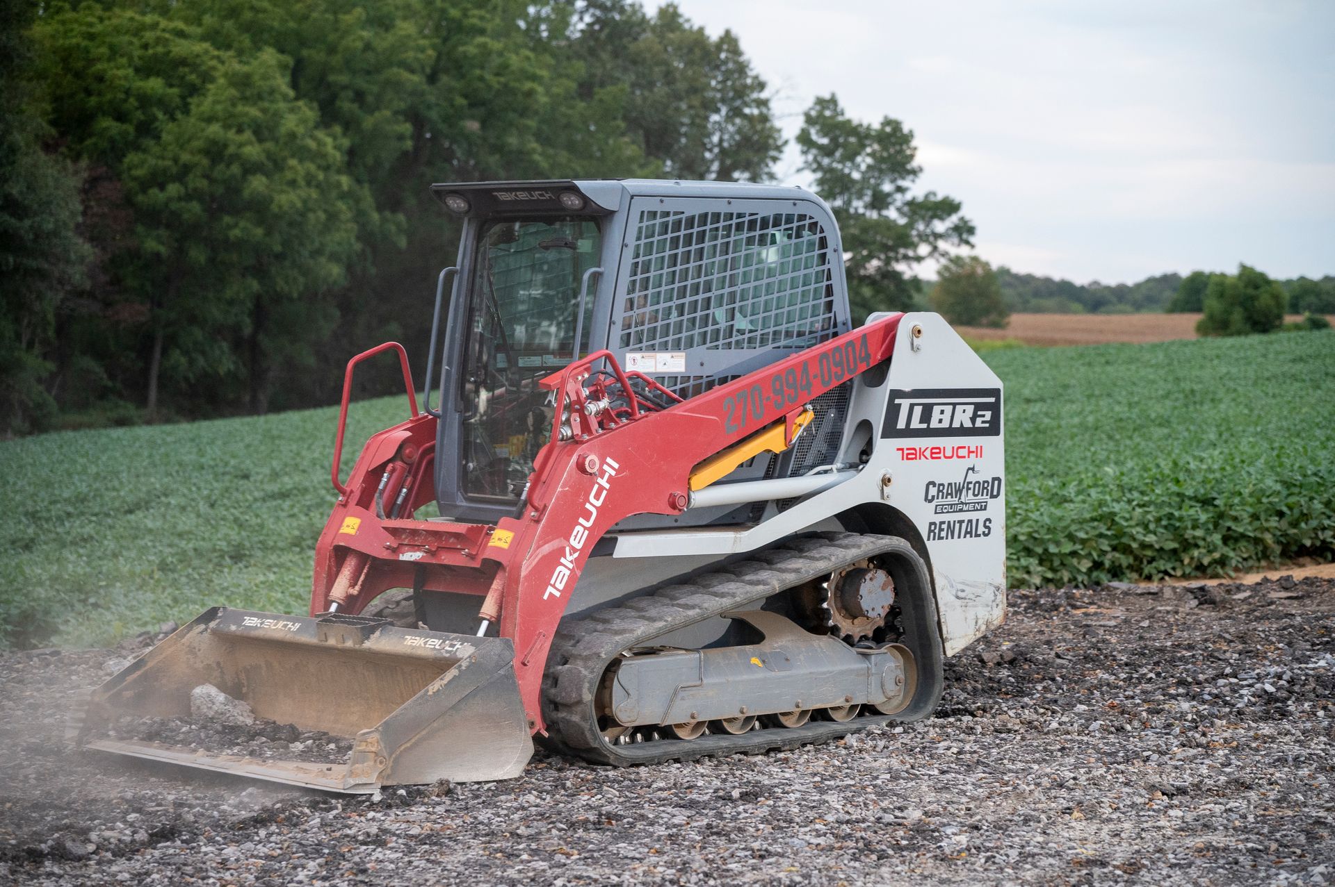 Tracked skid steer, red and gray, scoops gravel in a field.