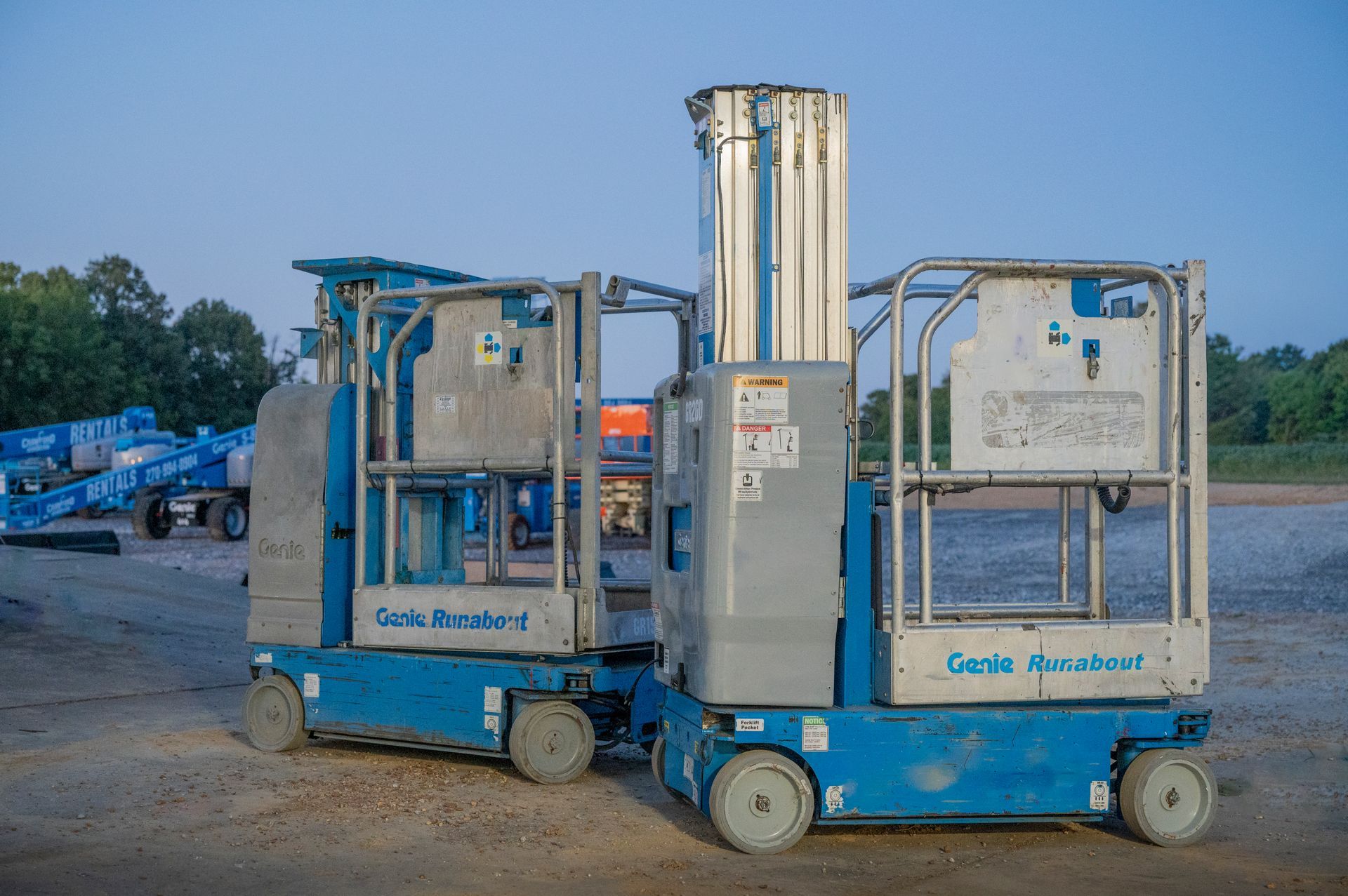 Two blue and gray aerial work platforms on gravel.