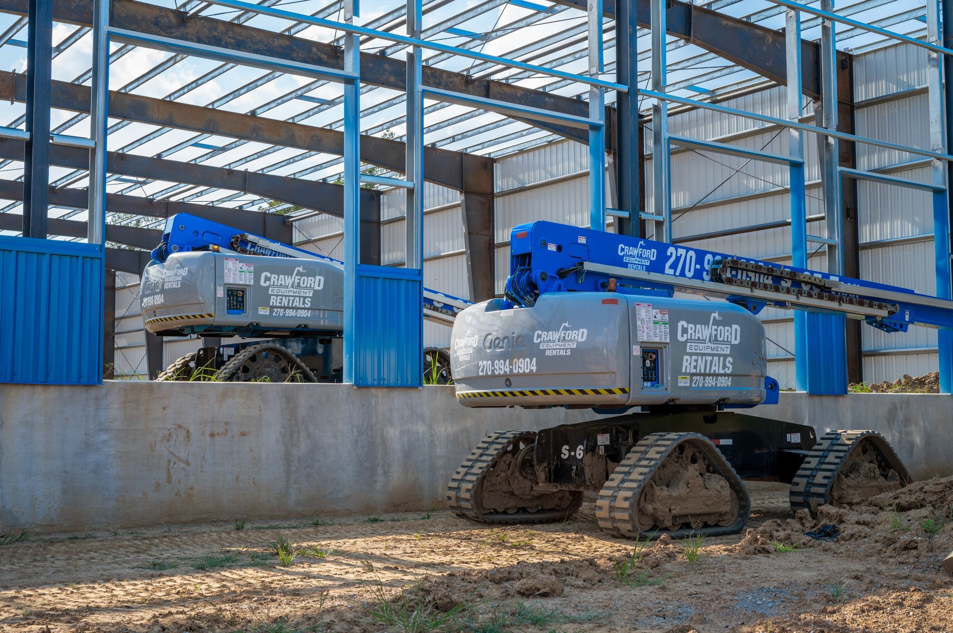 Two blue and gray boom lifts on tracks at a construction site with steel framing.