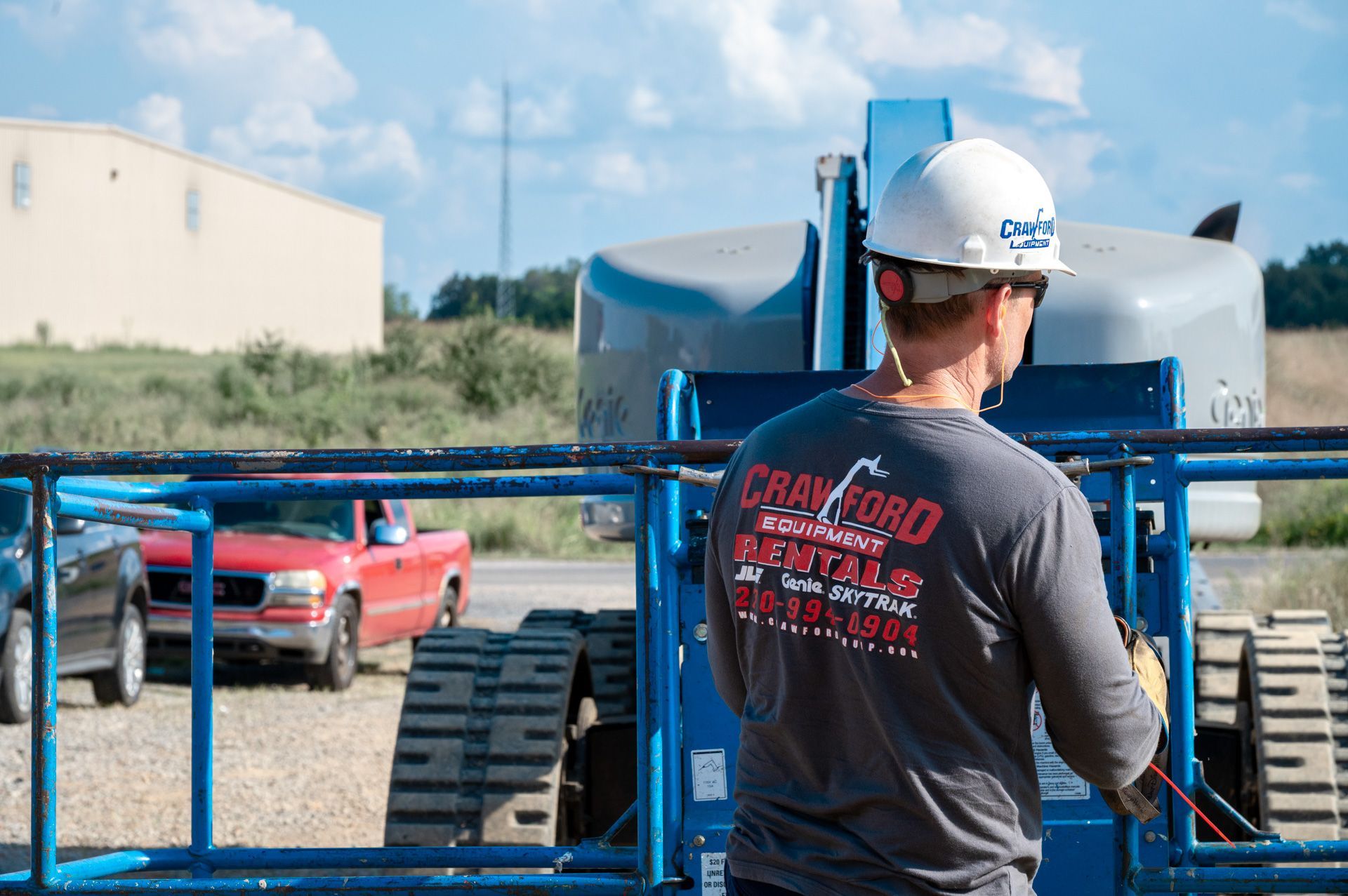 Construction worker in hard hat operates a blue lift with red truck in background.