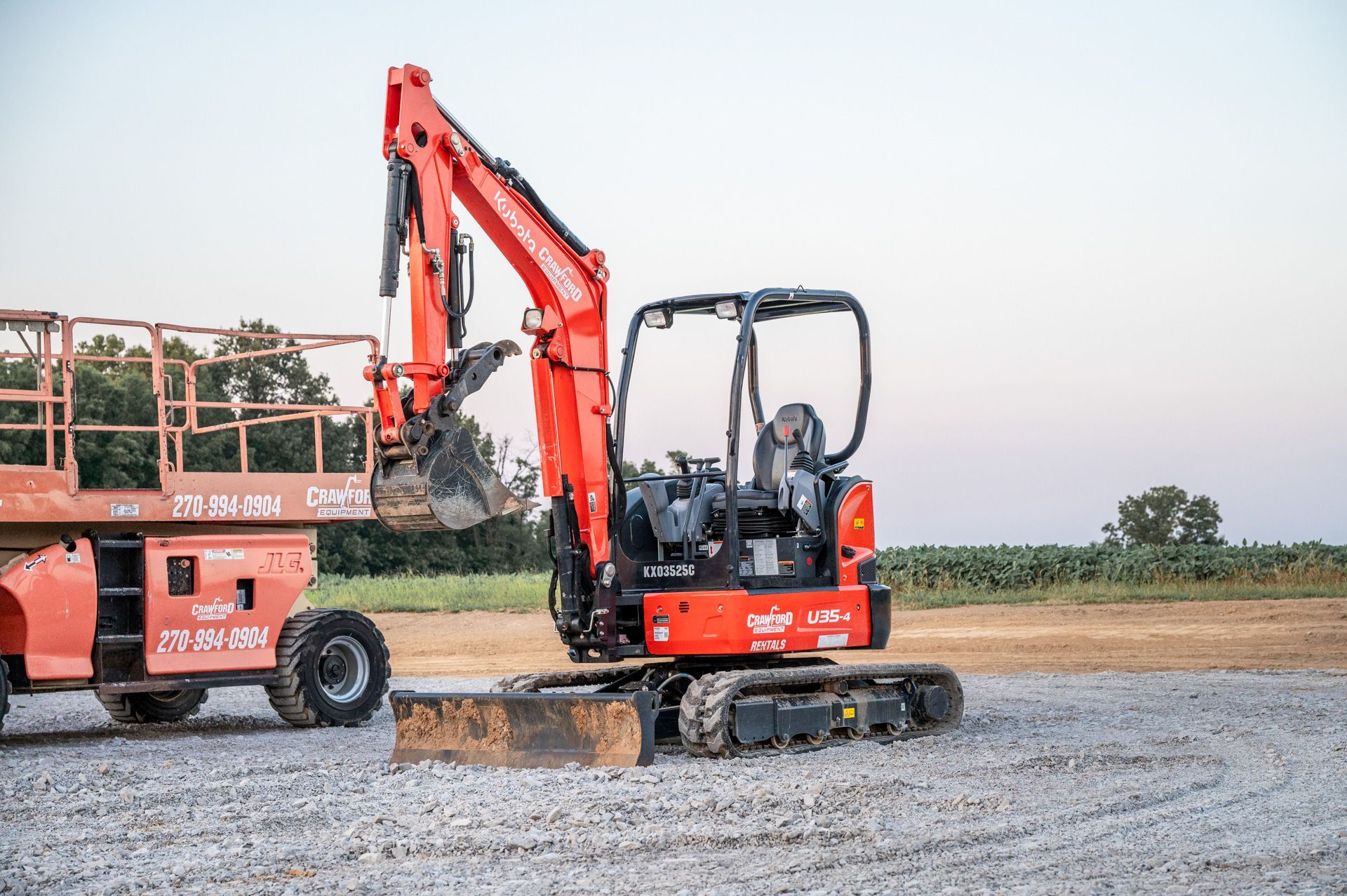 Orange Kubota excavator, ready for work, next to an orange lift. Gray gravel ground in field.