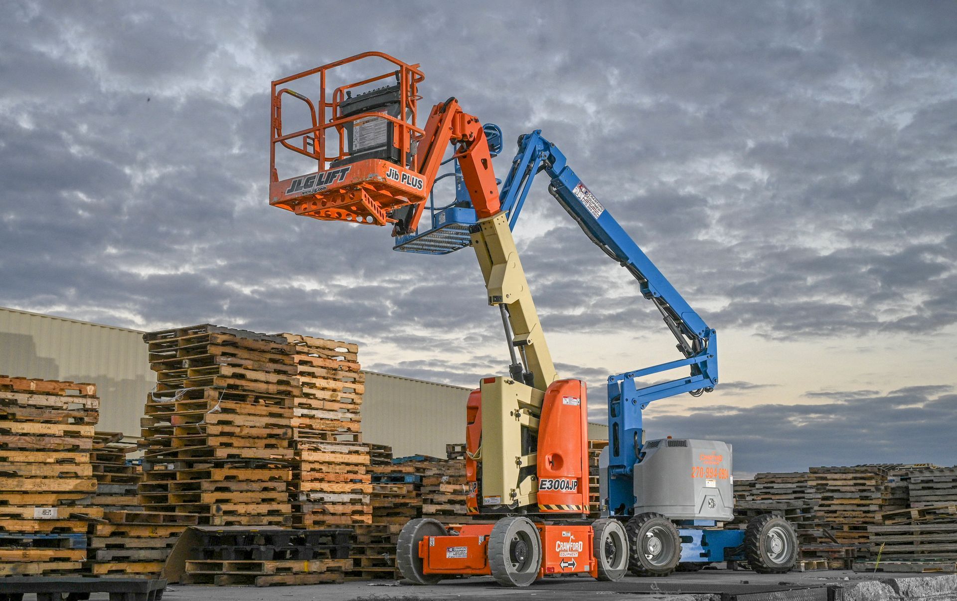 Orange and blue boom lift extending towards the sky near a stack of wooden pallets.