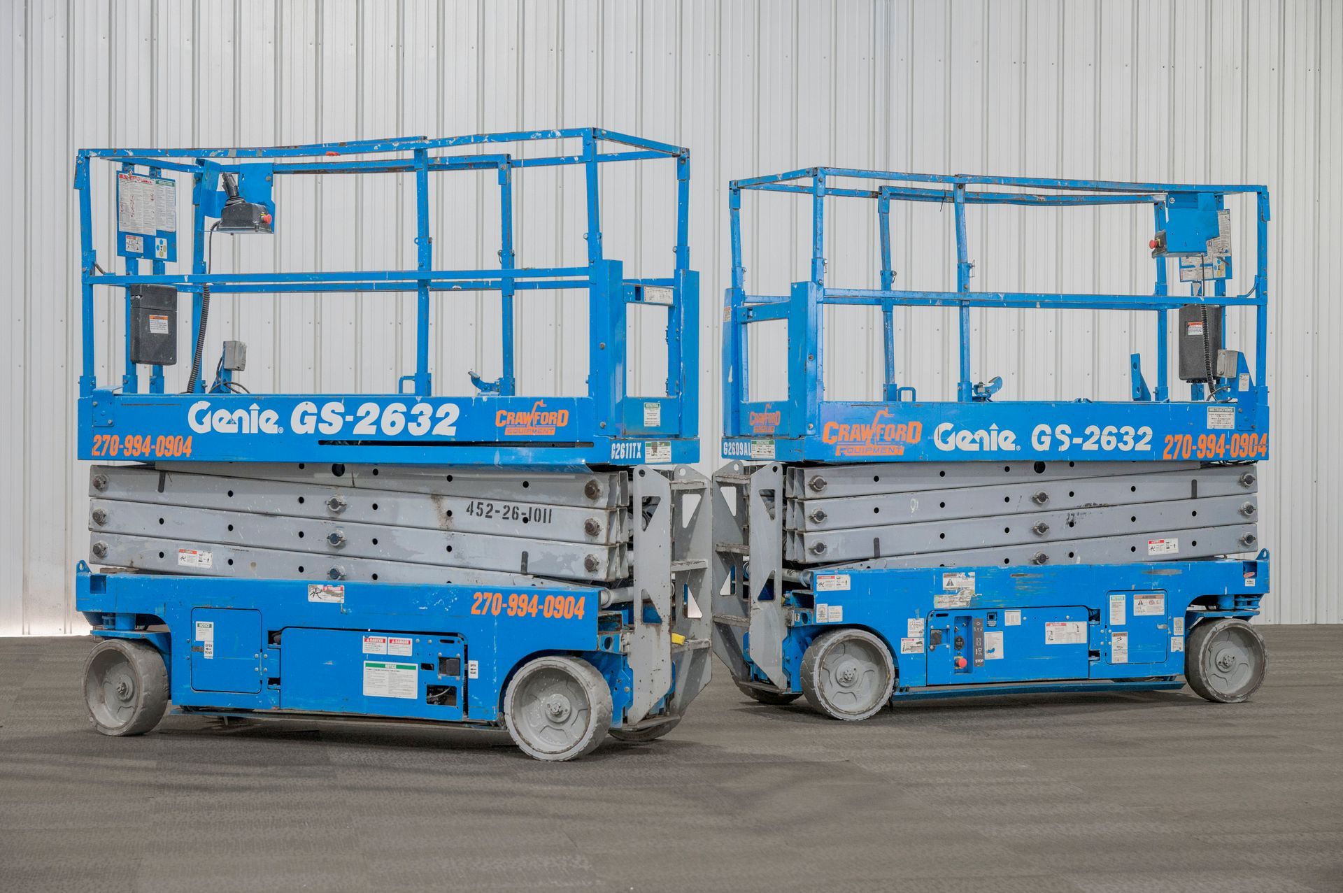 Two blue Genie GS-2632 scissor lifts side-by-side on a gray surface. They have blue railings and wheels.