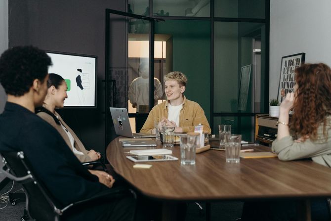 Group of people in a meeting around a table, discussing with laptops and a screen in an office.