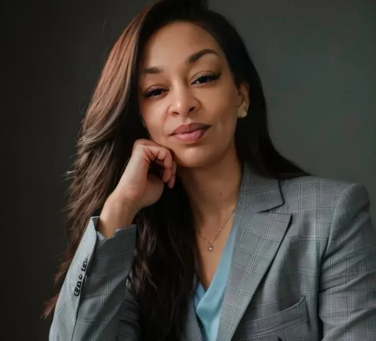 Woman with long brown hair, wearing a gray blazer, resting chin on hand, posing with a calm expression.