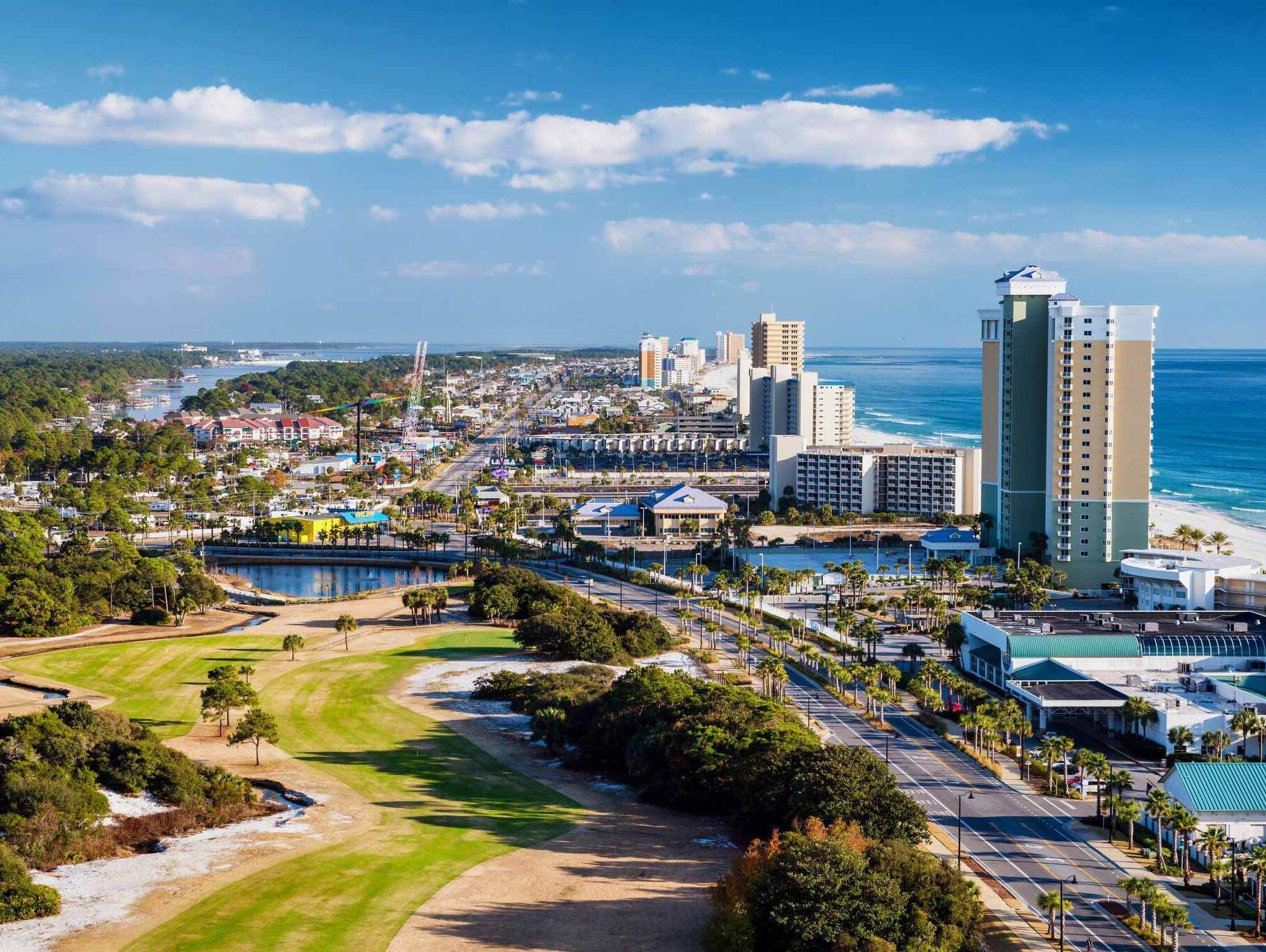 Aerial view of a coastal city with a beach, buildings, and a golf course under a blue sky.