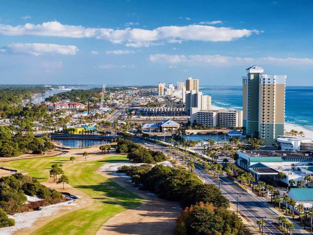 Aerial view of a coastal city with a beach, buildings, and a golf course under a blue sky.