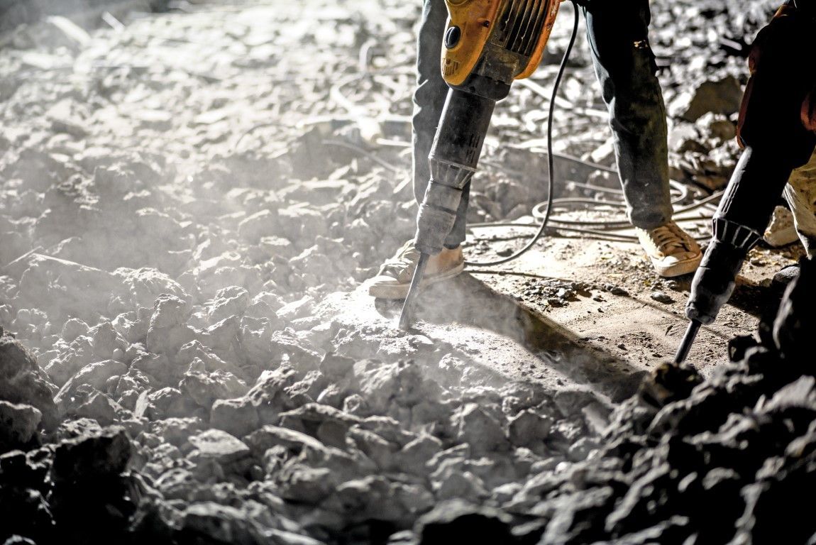 Two workers using jackhammers on rocky ground, surrounded by debris and dust, creating a rugged, intense industrial scene.