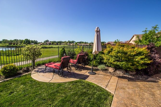 Two red lounge chairs and a closed umbrella on a sunlit patio with green grass, shrubs.