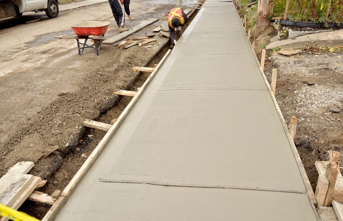 A worker in a reflective vest smooths a freshly poured concrete sidewalk. Nearby, a red wheelbarrow sits on a dirt road.