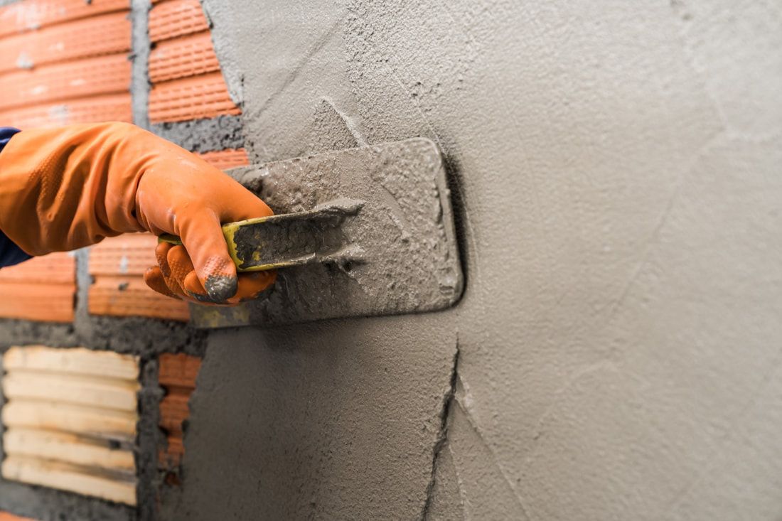 A hand in an orange glove applies smooth, wet cement to a brick wall using a trowel.