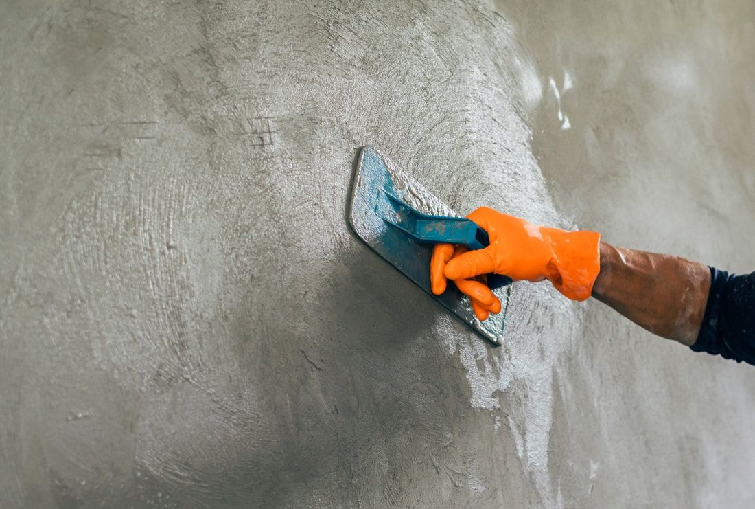 A person wearing an orange glove uses a blue trowel to smooth a gray concrete wall.
