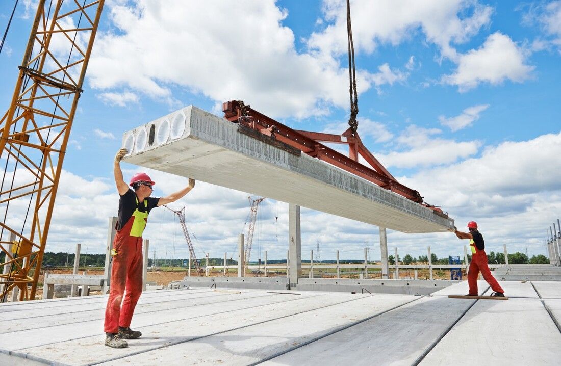 Two construction workers in red overalls guide a large concrete slab being lifted by a crane.