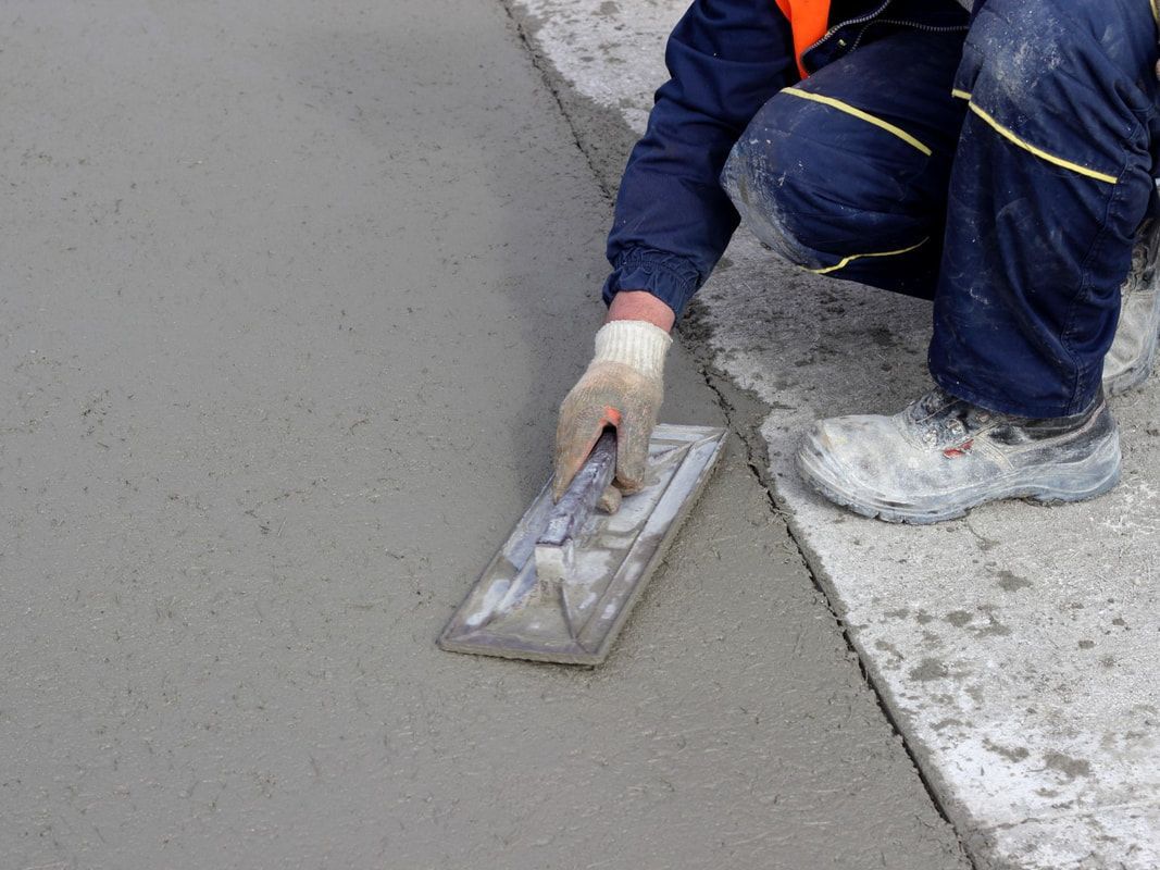 A person wearing work gloves is kneeling on a concrete surface, smoothing wet cement with a trowel.