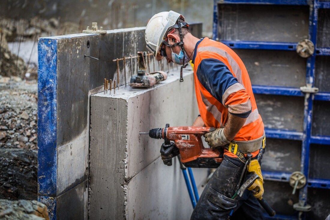 Worker in an orange safety vest and helmet drills into a concrete wall at a construction site, conveying focus and precision.