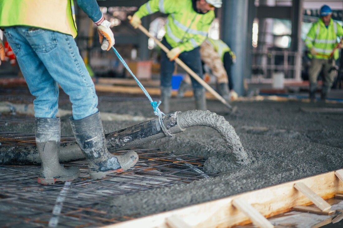 Construction workers in neon safety vests pour concrete onto a steel mesh foundation. 