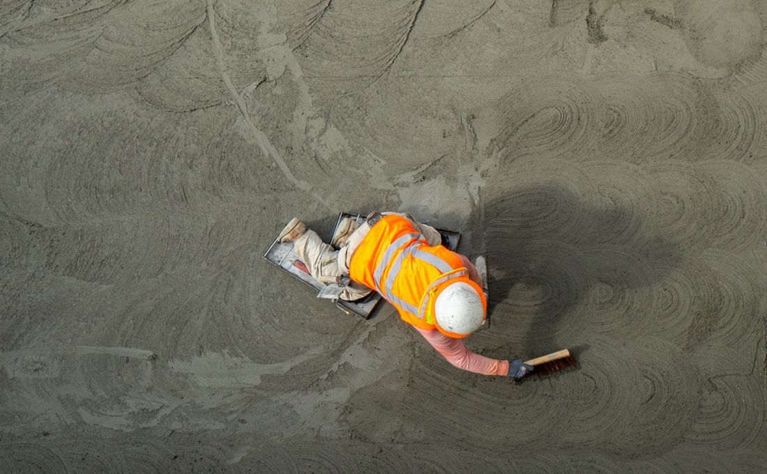 Worker in high-visibility orange vest and hard hat smoothing wet concrete with a trowel.