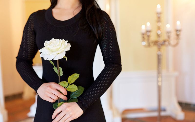 Una mujer vestida de negro sostiene una rosa blanca cerca de un candelabro encendido, probablemente en un funeral.