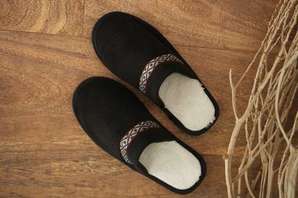 Un par de zapatillas negras sin cordones con forro de piel sintética blanca y una banda geométrica decorativa sobre una superficie de madera.