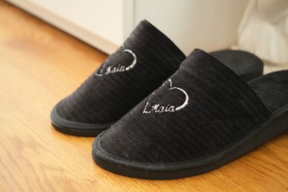 Un par de zapatillas negras texturizadas con un corazón blanco y un nombre bordados, sobre un suelo de madera.