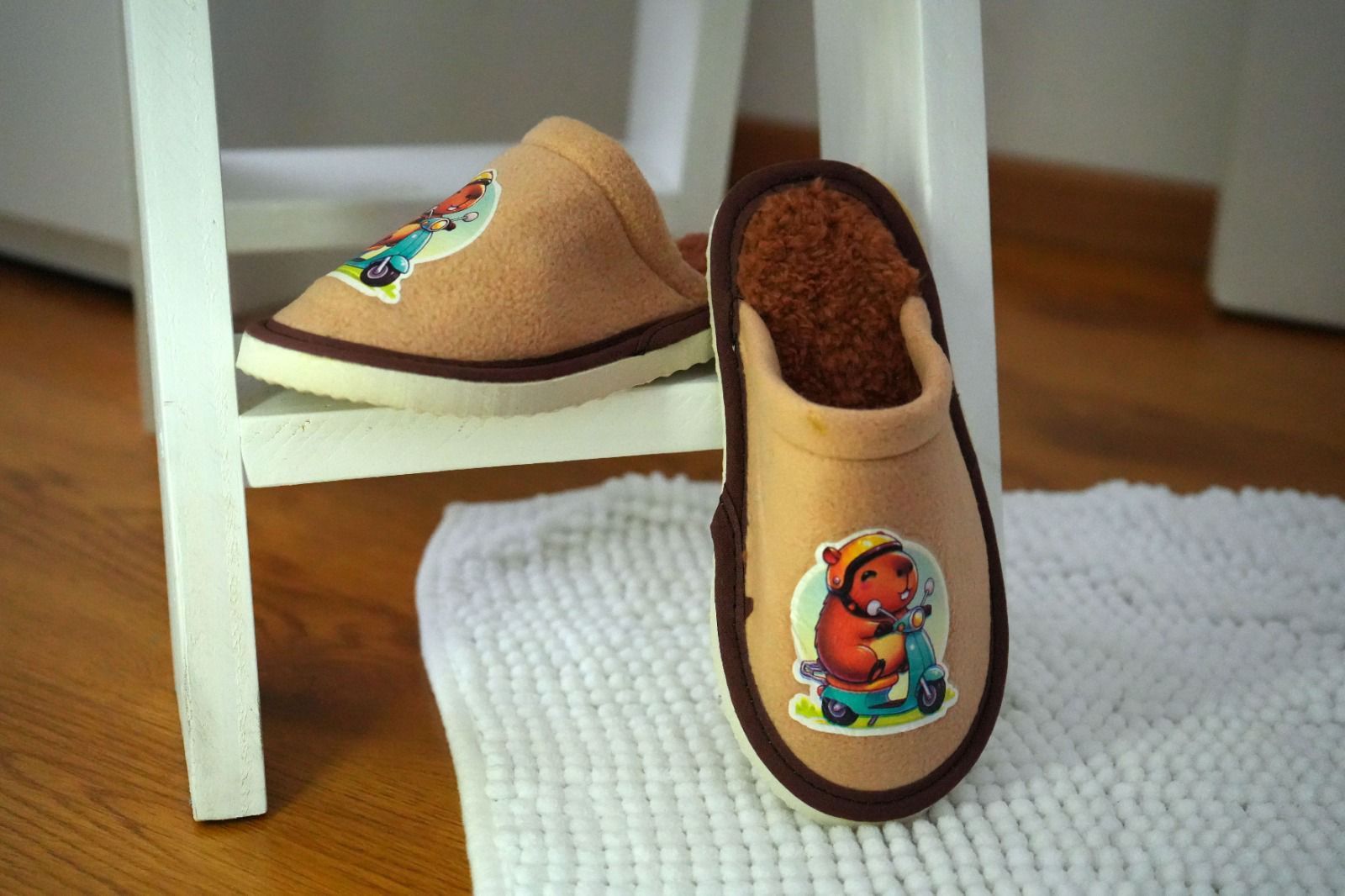 Zapatillas de fieltro color canela con ribete marrón y un animal ilustrado en un patinete; una descansa sobre una silla y la otra sobre una alfombra.