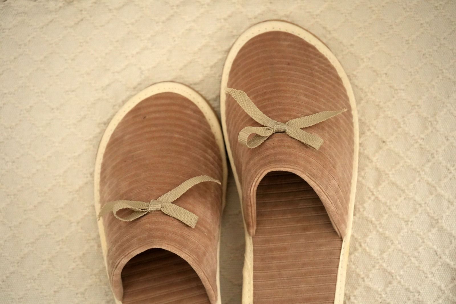 Cuatro zapatillas de casa en negro, rosa, gris y beige, cada una con un pequeño lazo, dispuestas una al lado de la otra sobre una alfombra texturizada.