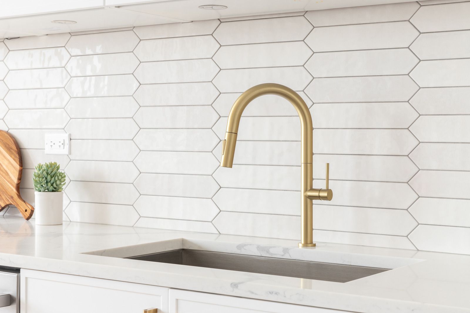Gold kitchen faucet over a stainless steel sink in a white kitchen with picket tile backsplash.