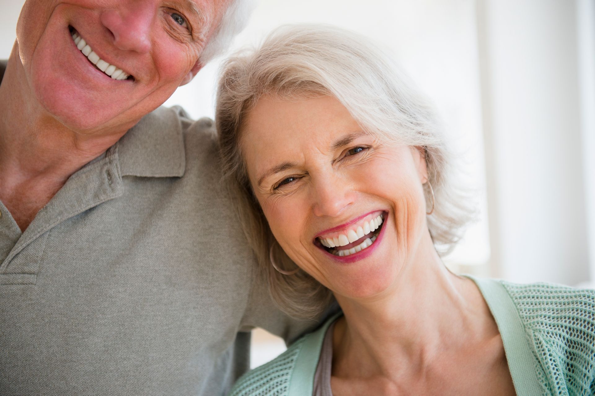 Coppia anziana sorridente, uomo con braccio intorno alla donna, entrambi con i capelli grigi, ambiente interno.
