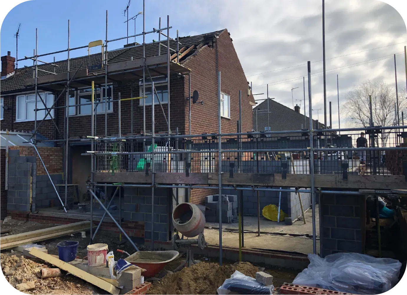 Construction site with scaffolding around a two-story brick house, visible foundation and cement mixer.
