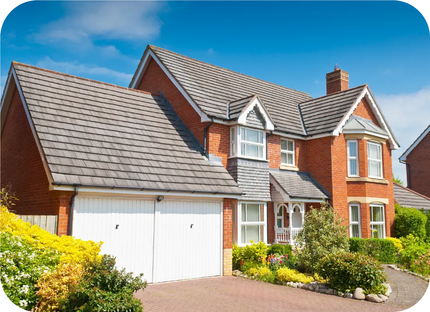 Brick house with gray roof, white garage doors, and lush landscaping against a blue sky.