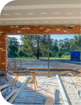 Interior view of a building under construction, looking out at a treed landscape. Brick, wood, and unfinished ceiling are visible.