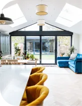 Bright kitchen and living space with a large window. Yellow chairs at a white island. Blue velvet sofa.