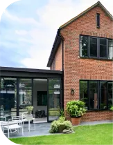 Brick house with black-framed windows and a glass-enclosed extension, viewed from a green lawn with patio furniture.