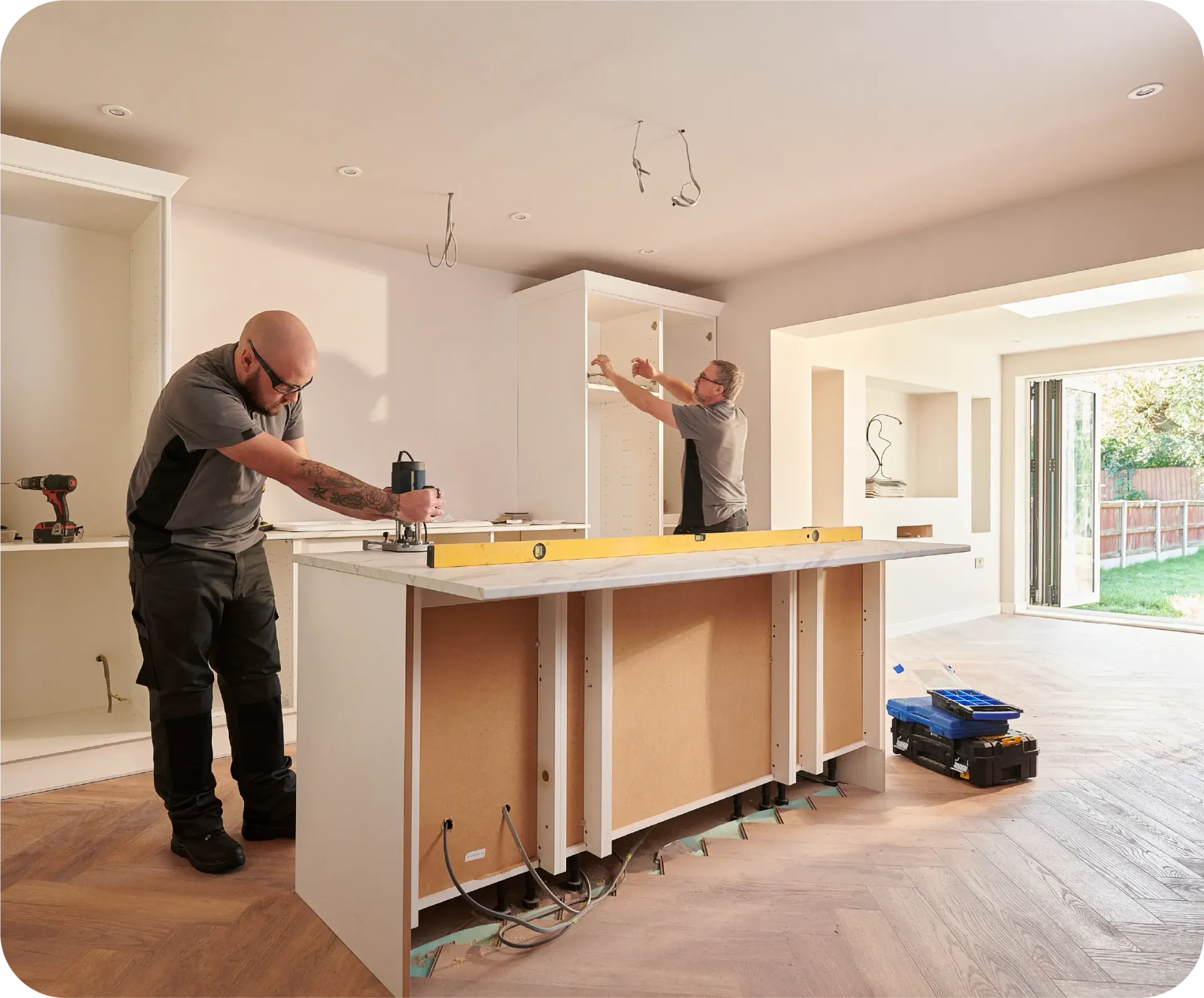 Two people installing a kitchen island in a bright, modern room. One uses a power tool, the other works on upper cabinets.