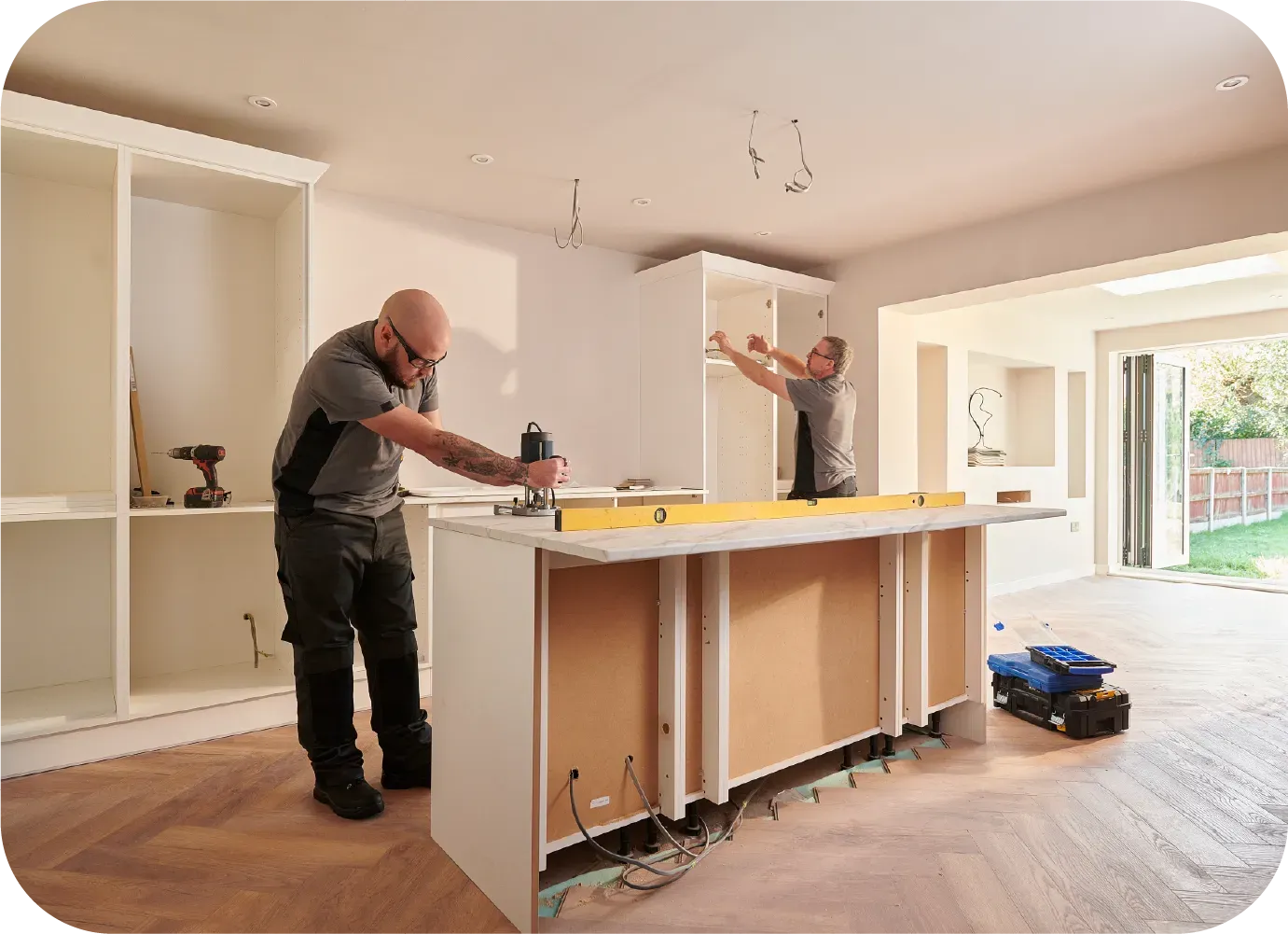 Two workers installing white kitchen cabinets in a room with wood flooring. One uses a power tool.